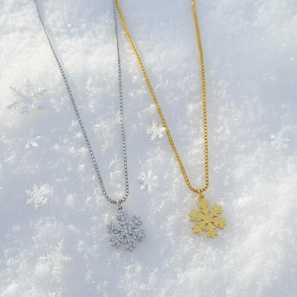 Two snowflake pendant necklaces on delicate chains, one silver-toned and one gold-toned, displayed on a soft pink fabric background.