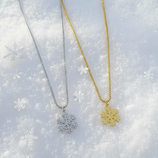 Two snowflake pendant necklaces on delicate chains, one silver-toned and one gold-toned, displayed on a soft pink fabric background.