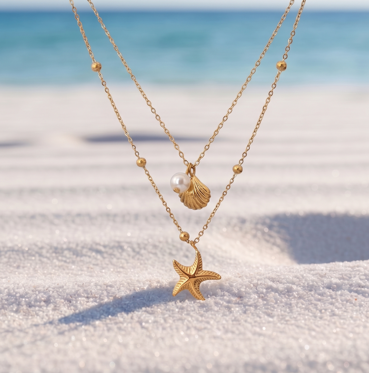 Two delicate gold chain necklaces with small bead accents are displayed on white sand at the beach. One pendant is a textured gold starfish, and the other features a gold scallop shape