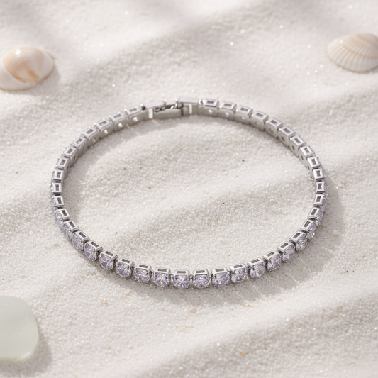 A silver tennis bracelet with clear cubic zirconia stones, displayed on a textured white surface with a stone prop in the corner.